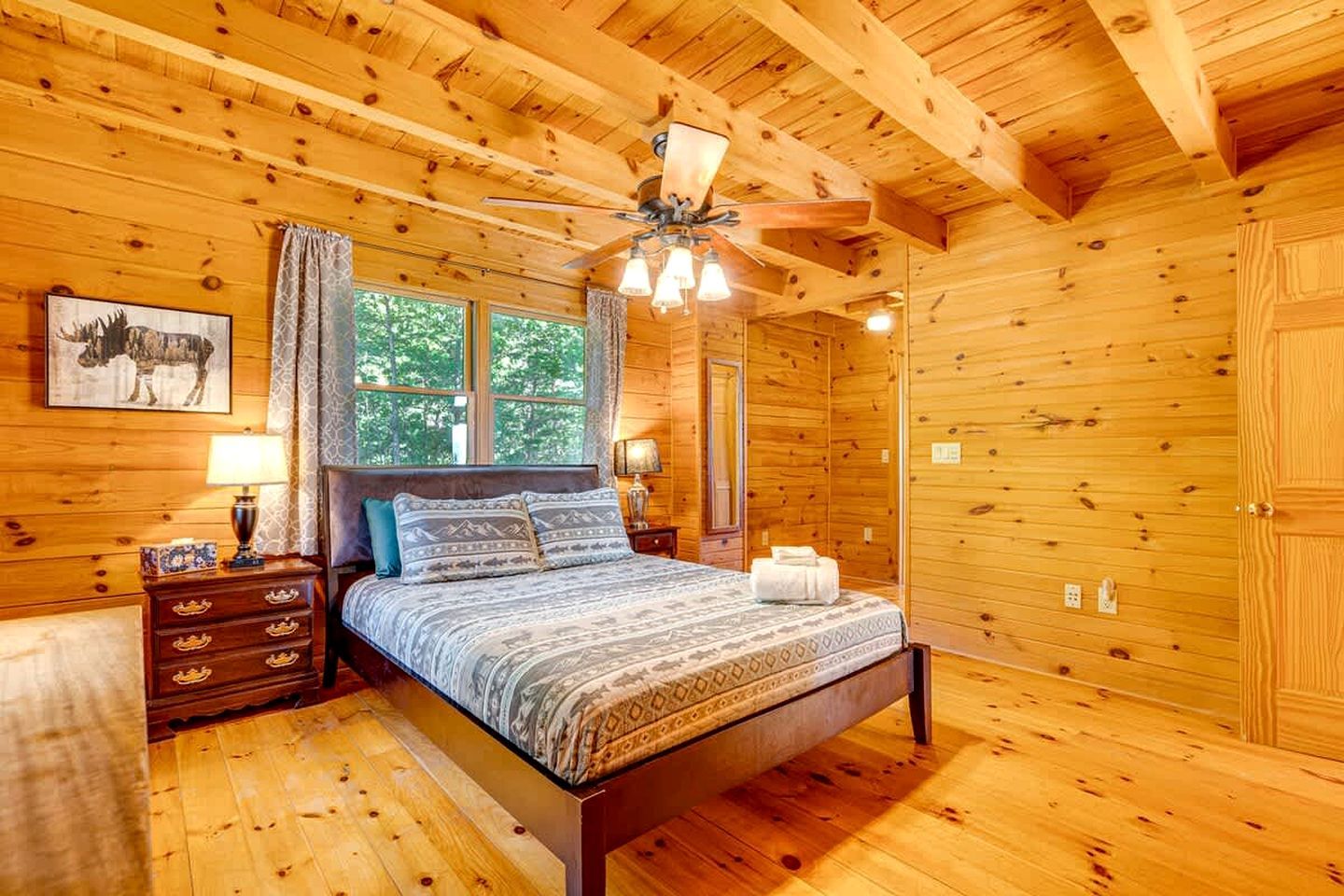 Un dormitorio de cabaña de madera con cama, mesitas de noche, ventilador de techo y ventana con cortinas.