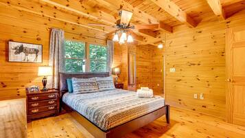 Un dormitorio de cabaña de madera con cama, mesitas de noche, ventilador de techo y ventana con cortinas.