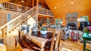 A spacious wooden interior with a central dining area, a fireplace, and a staircase leading to an upper level.