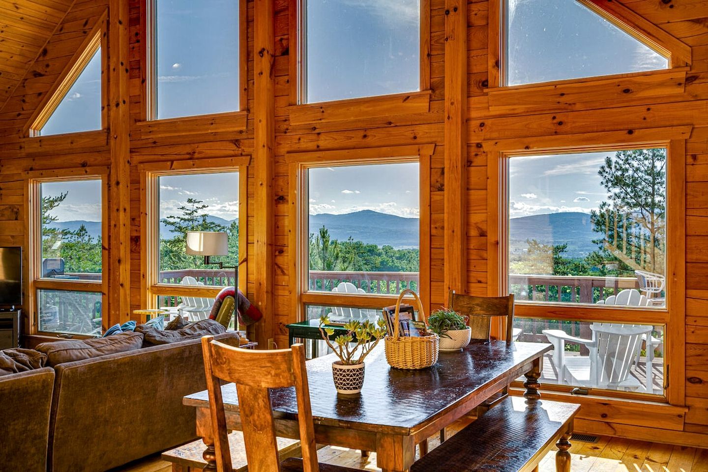 A wooden interior with a dining table, chairs, and a sofa. Large windows offer views of greenery and mountains.