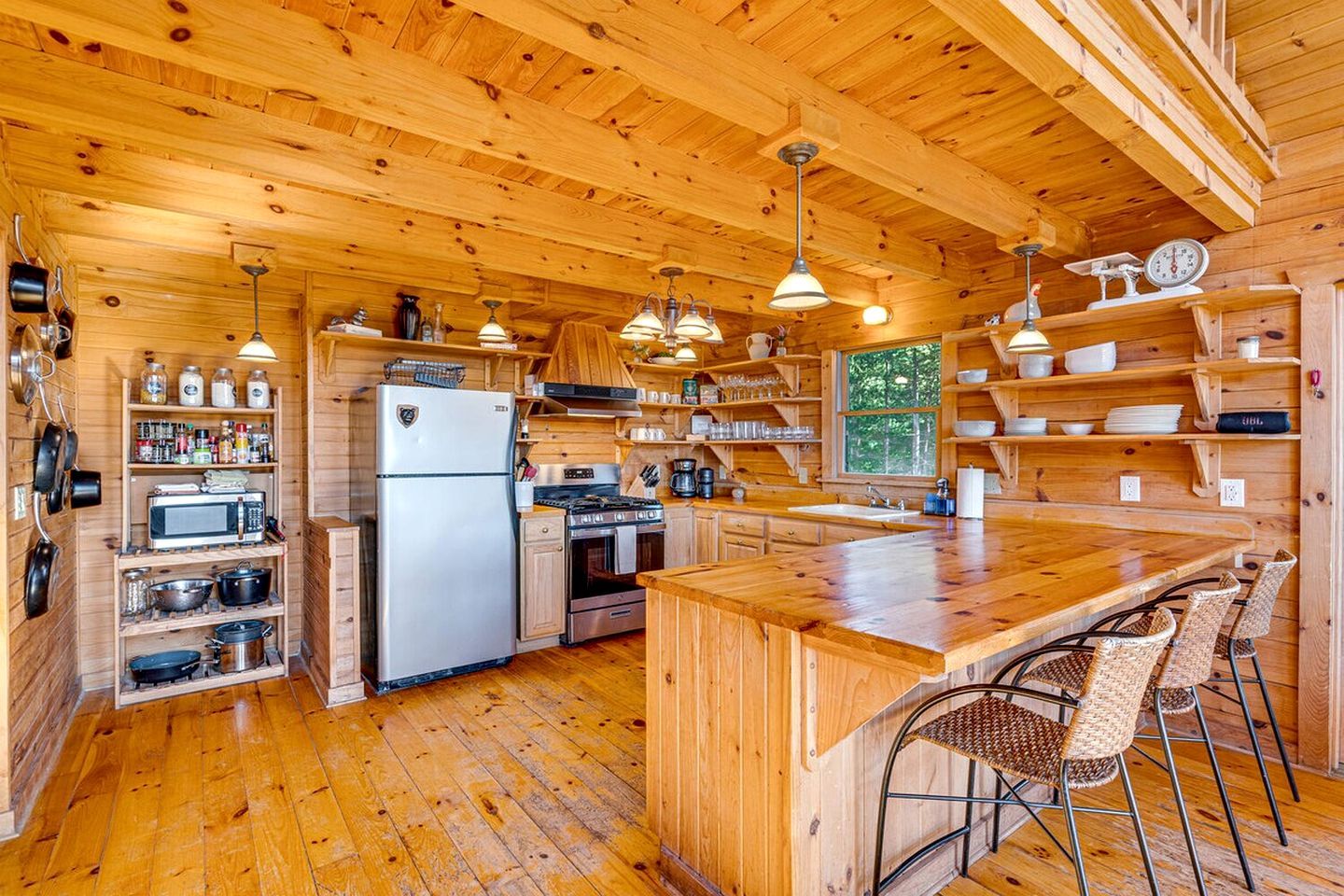 A wooden cabin interior with a kitchen featuring a refrigerator, stove, oven, and various kitchen utensils.