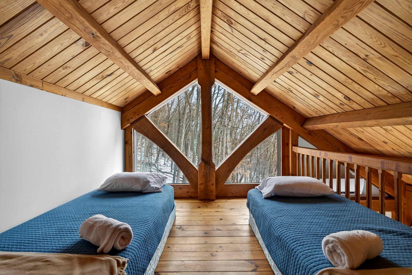 A room with a sloped wooden ceiling, two beds with blue bedding, and a view of trees through a large window.