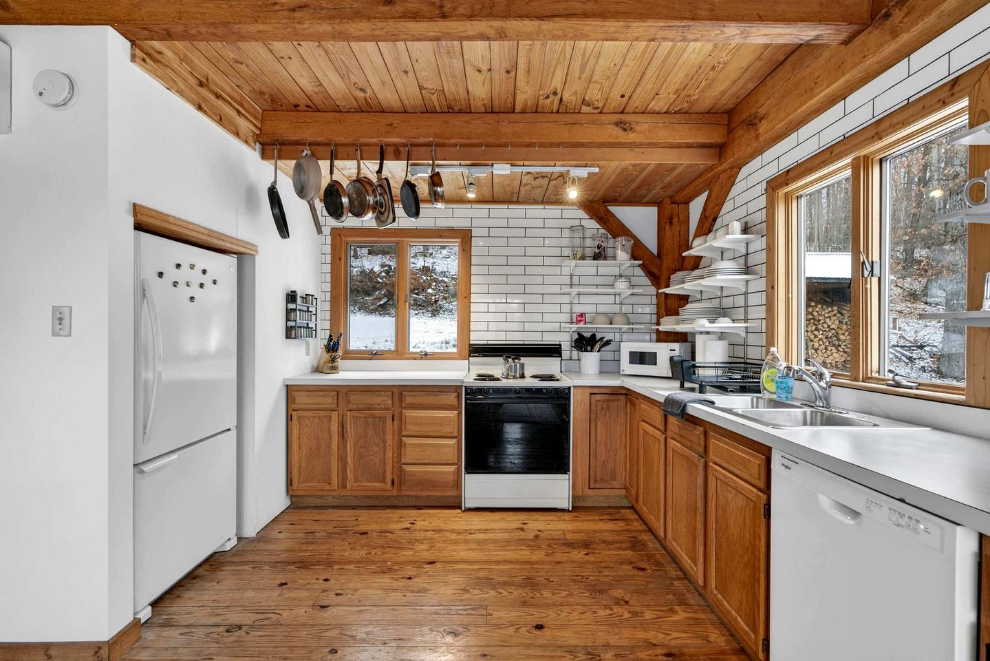 A kitchen with wooden cabinets, a white refrigerator, a dishwasher, and a window with a view of trees.