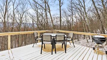 A wooden deck with a table and chairs, a grill, and a view of trees and a body of water.