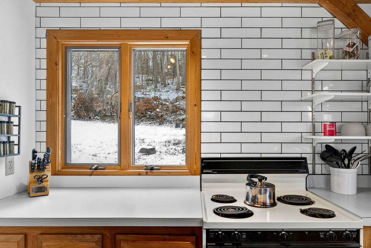 A kitchen with a double window, white tiled backsplash, and a stove with a kettle.