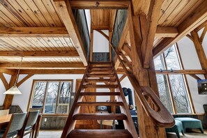 A wooden staircase leading to an upper level, with a dining area below featuring a table and chairs. - Charming Post-and-Beam Mountain Cabin with Hot Tub Near Berkeley Springs | Great Cacapon, WV (Great Cacapon)