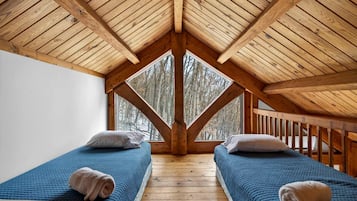 A room with a sloped wooden ceiling, two beds with blue bedding, and a view of trees through a large window.
