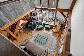 Uma ampla sala de estar no estilo loft, com teto alto, vigas de madeira e uma grande janela com vista para uma paisagem nevada.
