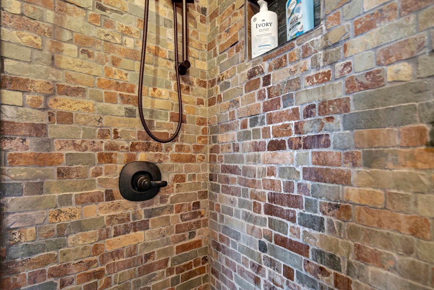 A shower with a brick wall, a handheld showerhead, and toiletries on a shelf.