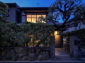 Una casa japonesa tradicional amb entrada de pedra, envoltada d'arbres de pi i un jardí ben cuidat.
