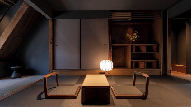 Un intérieur moderne au design minimaliste, comprenant une table centrale avec deux chaises, une lampe et une étagère le long d’un mur sombre.