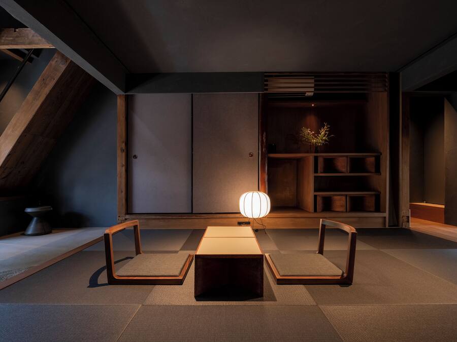 Um interior moderno com design minimalista, apresentando uma mesa central com duas cadeiras, uma luminária e uma estante em frente a uma parede escura.