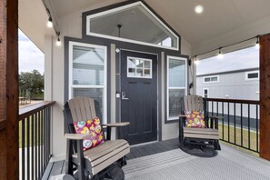A covered porch with two rocking chairs, a dark door, and a view of a building in the background. - Relaxing Tiny Home Retreat with Resort Amenities Near Fredericksburg, TX (Fredericksburg)