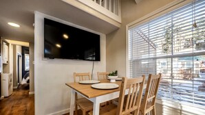 A dining area with a TV mounted on the wall, a table with chairs, and a window with blinds. - Quaint Tiny House in Woodland Park, Colorado near Garden of the Gods (Woodland Park)