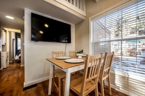 Un comedor con un televisor montado en la pared, una mesa con sillas y un ventanal con persianas.