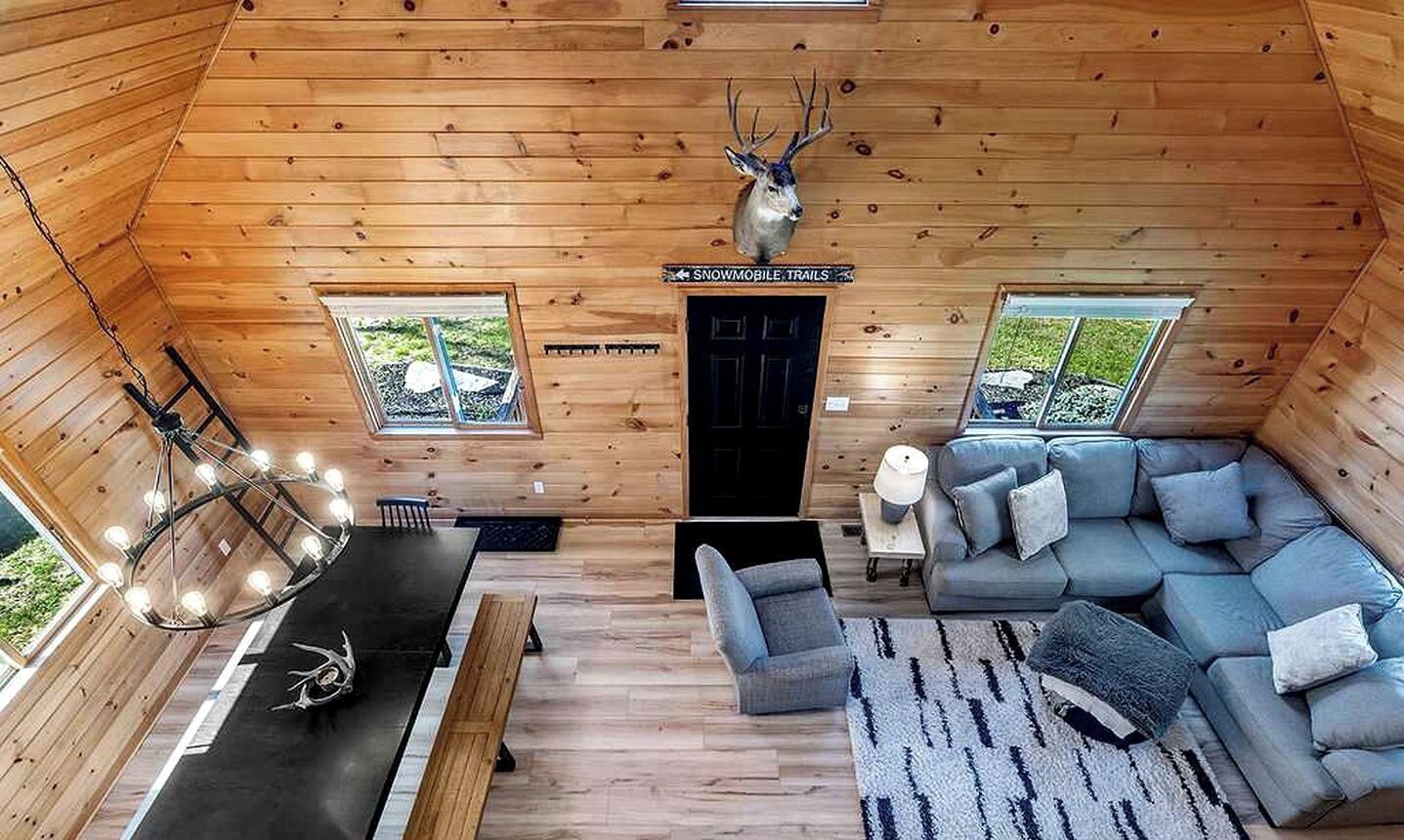 A cabin interior with a wooden ceiling, a mounted deer head, a sofa, a dining table, and a sign for 'SNOWMOBILE TRAILS'.