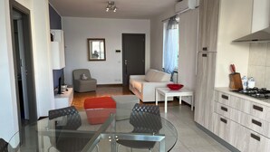 A modern apartment with a glass dining table, a sofa, a red ottoman, a kitchen with a stove, and a window with curtains. - Lakefront Escape | Private Terrace & Lake Access (Predore)