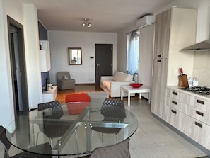 A modern apartment with a glass dining table, a sofa, a red ottoman, a kitchen with a stove, and a window with curtains. - Lakefront Escape | Private Terrace & Lake Access (Predore)