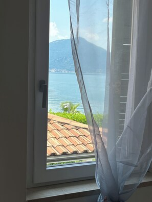 Una ventana con vista a un lago y montañas, un techo de tejas y una cortina blanca.
