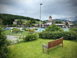 Blick auf einen See mit einer Bank, Booten und Gebäuden im Hintergrund.