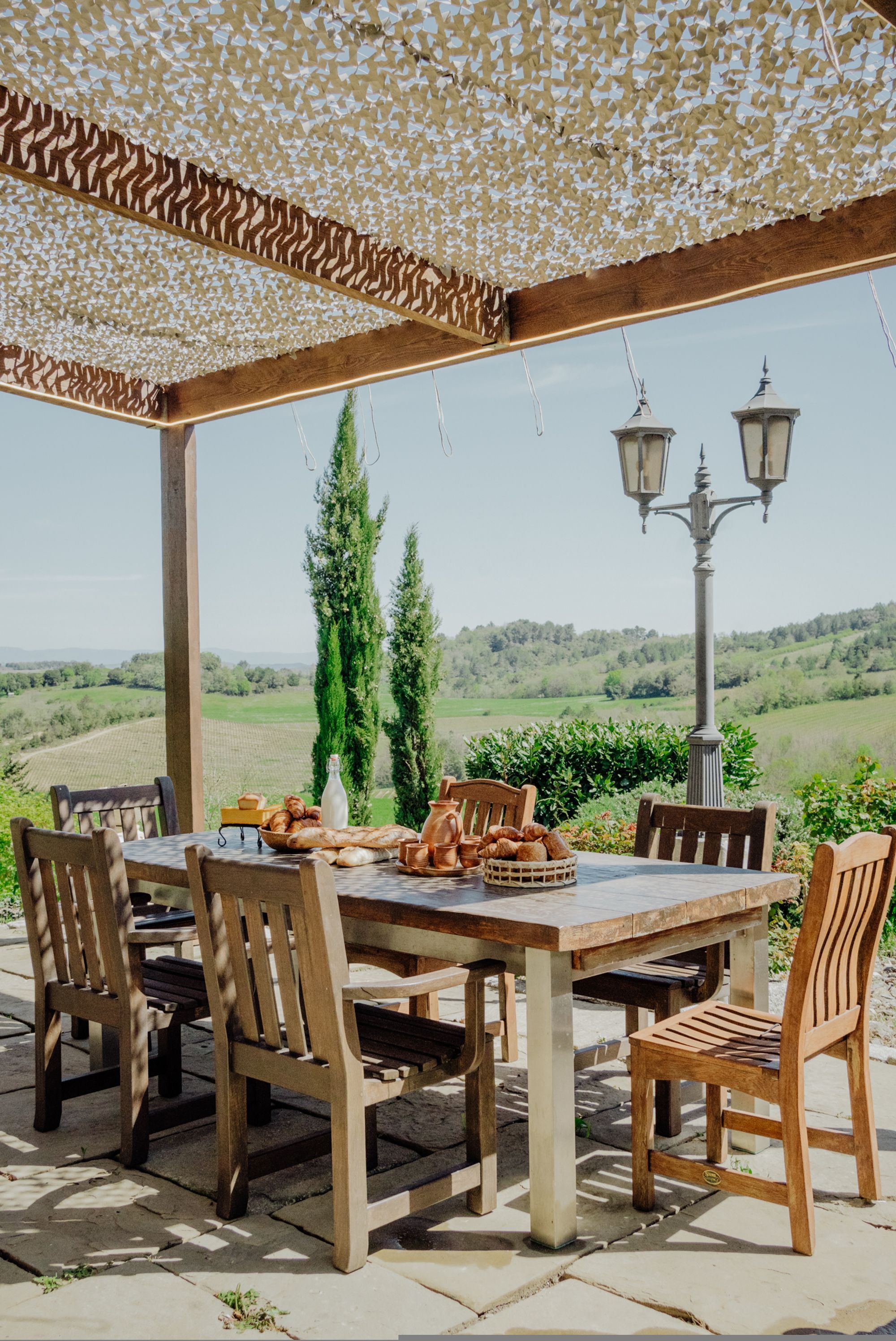 Un área de comedor al aire libre, cubierta, con una mesa dispuesta para seis personas, rodeada de sillas de madera, con vistas a un paisaje verde con colinas.