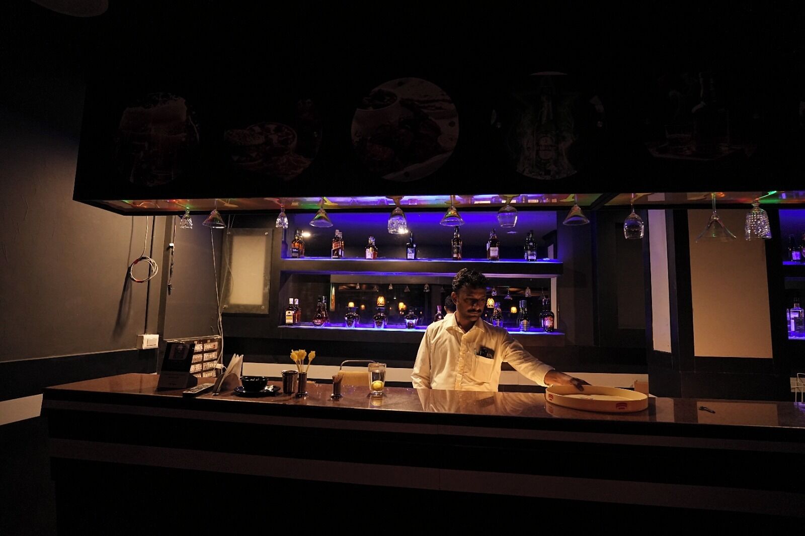 Zona de bar con un cantinero, estantes con botellas y un panel de menú.