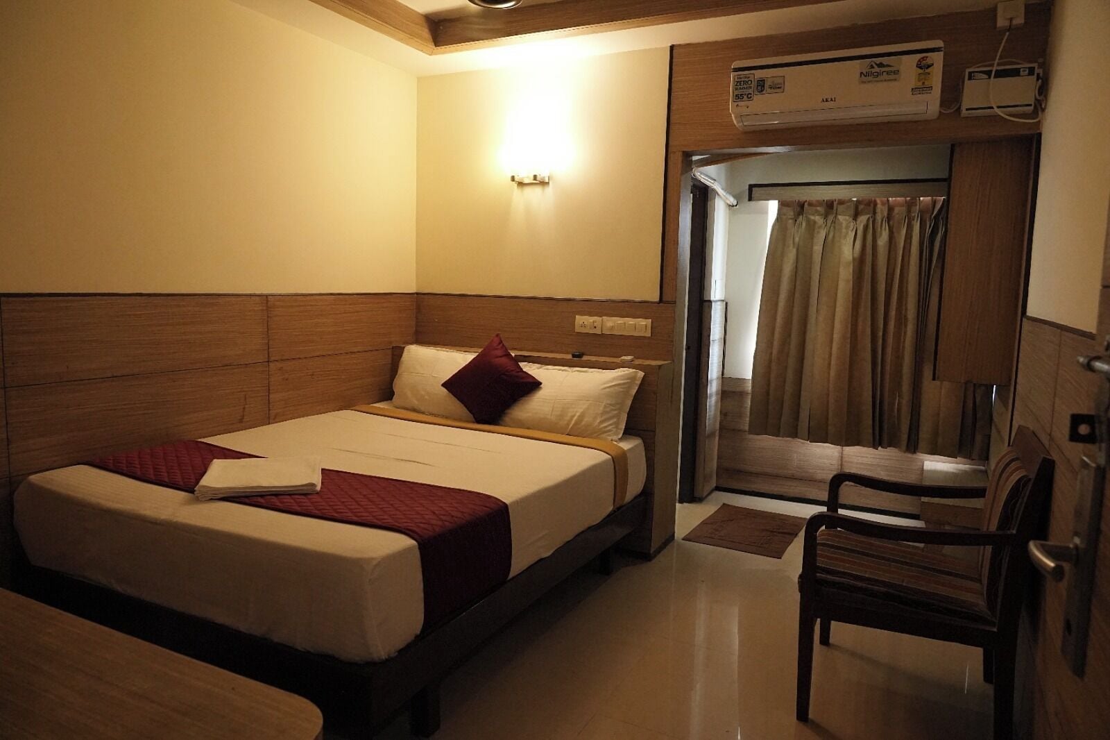 A hotel room with a bed, a chair, a wall-mounted air conditioner, and a curtain.