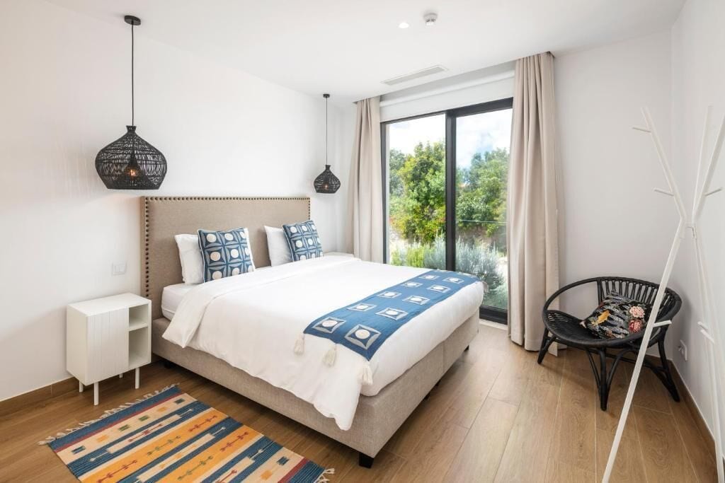 Quarto com cama, mesa de cabeceira, abajur, cadeira e vista para o verde lá fora.