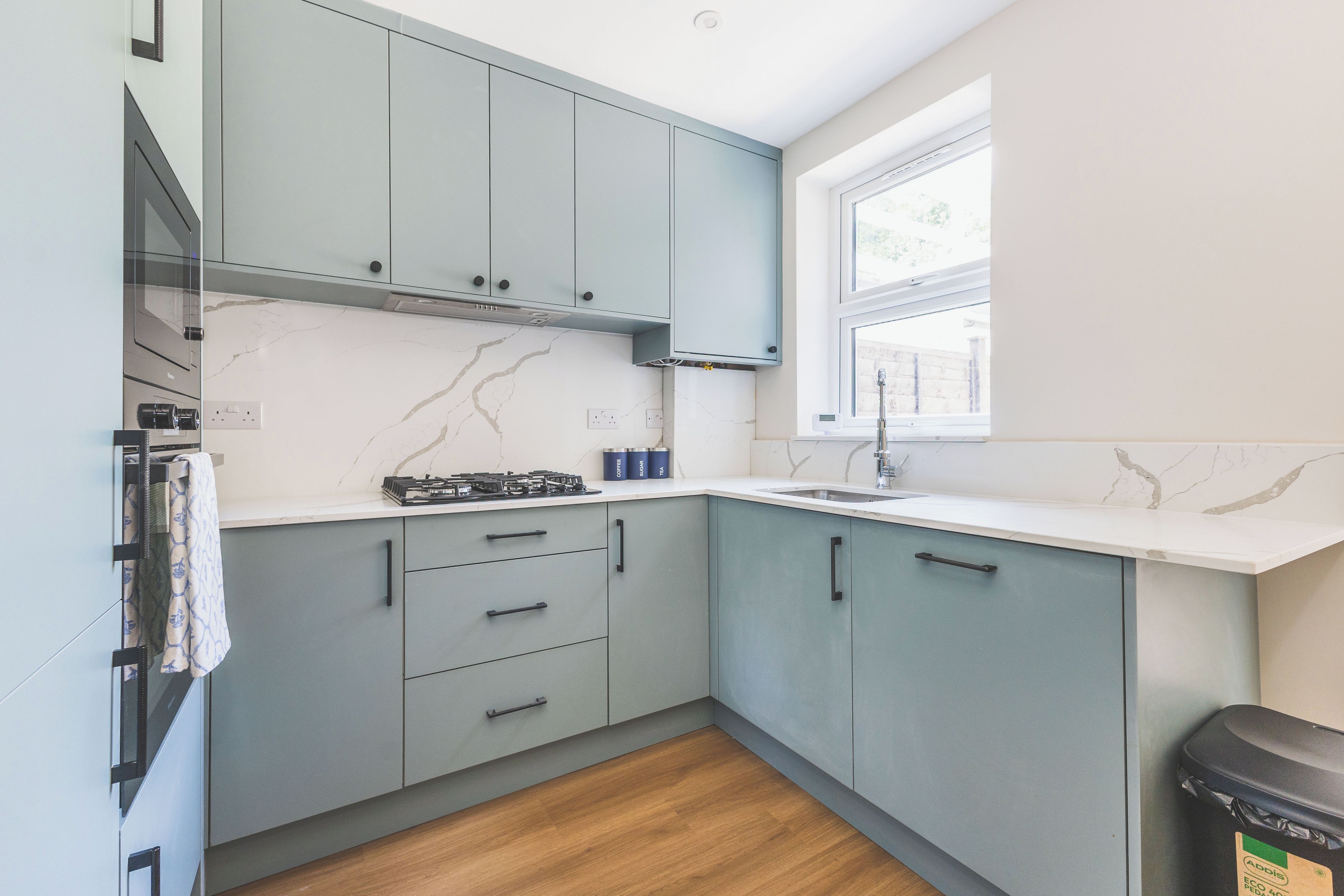 A modern kitchen with grey cabinets, a white countertop, and a window providing natural light.