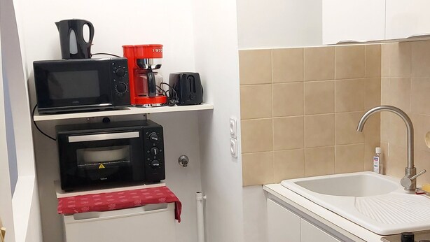 A compact kitchen with a microwave, toaster, and oven on a white countertop, a sink with a faucet, and beige tiled walls. - A charming studio on the French Riviera, 25 minutes from Nice, nestled between the sea and the mountains. (L'Escarène)