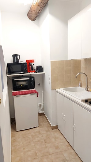 Cocina compacta con microondas, tostadora y horno sobre una encimera blanca, un lavabo con grifo y paredes de azulejos color beige.