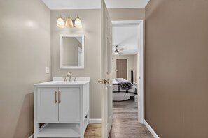 Ein Badezimmer mit einem weißen Waschtisch, einem Spiegel und einer Tür zu einem Schlafzimmer mit Bett und Deckenventilator.