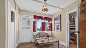 Ein Zimmer mit Holzboden, einer weißen Tür, einem Fenster mit roten Vorhängen, einer Couch, einem Tisch mit Blumenvase und einem Regal mit Handtüchern.