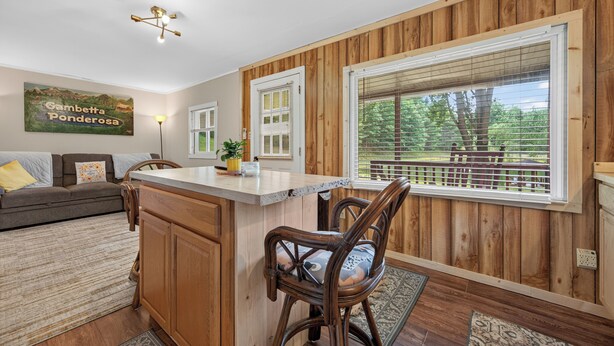 A kitchen with wooden paneling, a central island with chairs, a dining area with a sofa, and a large window with blinds. - Cozy lakeside cabin with peaceful views, a covered porch, fire pit, and private rural getaway. (Galax)