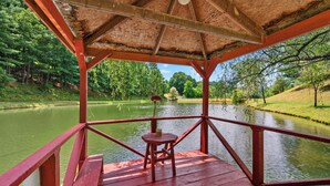A wooden deck with a red railing overlooking a calm river, surrounded by lush greenery. - Cozy lakeside cabin with peaceful views, a covered porch, fire pit, and private rural getaway. (Galax)