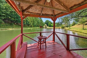 A wooden deck with a red railing overlooking a calm river, surrounded by lush greenery. - Cozy lakeside cabin with peaceful views, a covered porch, fire pit, and private rural getaway. (Galax)