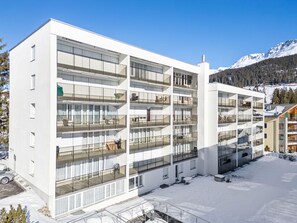 Un hotel de varios pisos con balcones, una zona de estacionamiento y una montaña nevada al fondo.