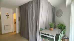 Een kamer met een eettafel, stoelen, een potplant, een klok aan de muur en een gordijn.