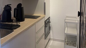 Een kleine keuken met witte kastjes, een roestvrijstalen afzuigkap, een koffiezetapparaat en een klein wit karretje met een mandje.
