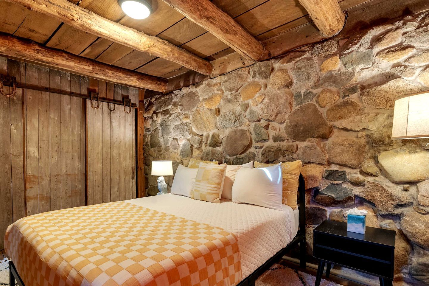 A bedroom with a stone wall, wooden ceiling beams, a bed with a checkered bedspread, and a nightstand with a lamp.