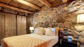 A bedroom with a stone wall, wooden ceiling beams, a bed with a checkered bedspread, and a nightstand with a lamp.