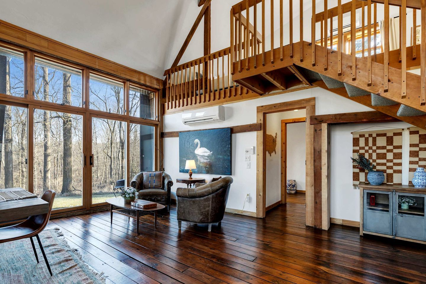A spacious living room with a wooden staircase, large windows, and a view of trees.