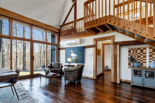 A spacious living room with a wooden staircase, large windows, and a view of trees.