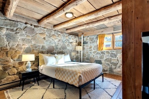 Quarto com paredes de pedra, uma cama, mesas de cabeceira e uma janela com cortinas.