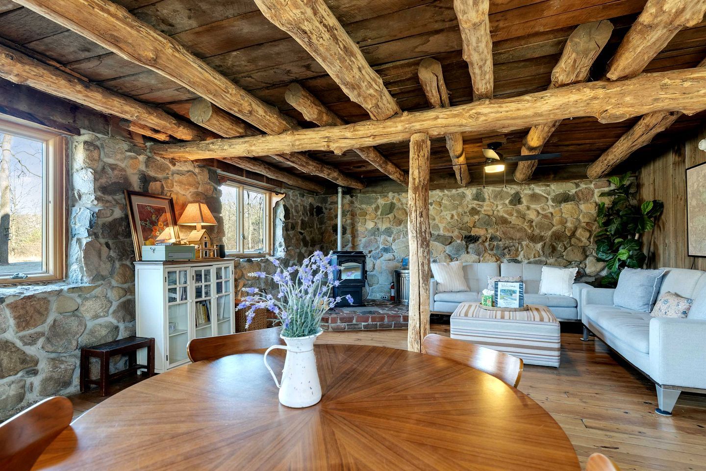 A rustic interior with a stone wall, wooden ceiling beams, a round wooden table, a sofa, and a fireplace.