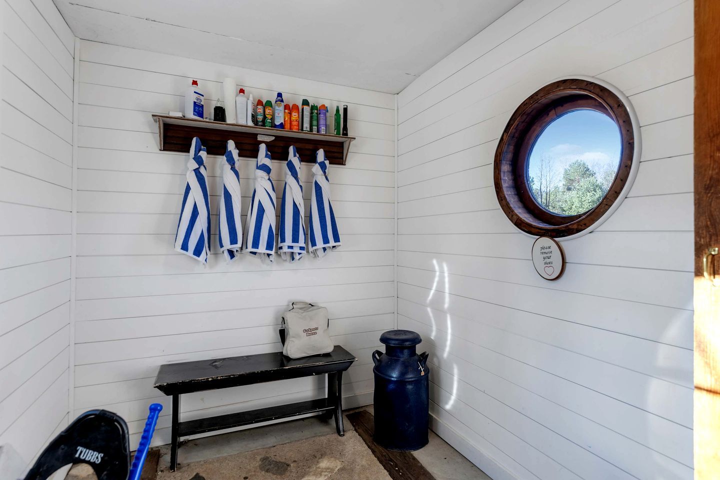 A small room with white walls, a wooden bench, a shelf with cleaning supplies, a window with a view, and a towel rack with striped towels.