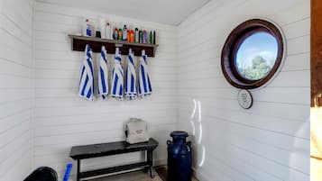 A small room with white walls, a wooden bench, a shelf with cleaning supplies, a window with a view, and a towel rack with striped towels.