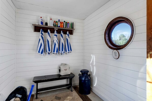 A small room with white walls, a wooden bench, a shelf with cleaning supplies, a window with a view, and a towel rack with striped towels.
