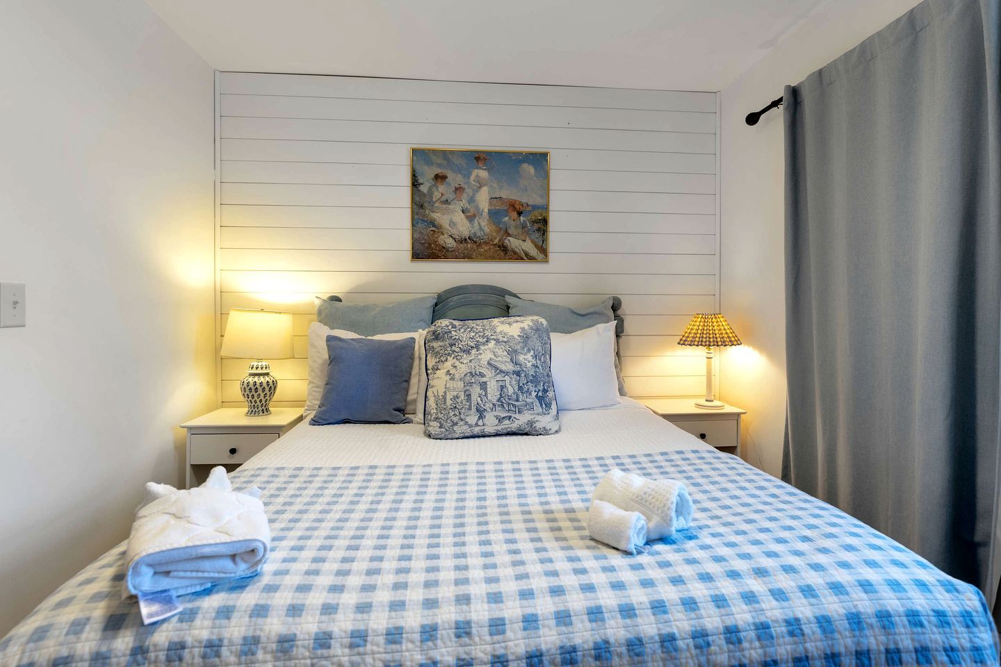 A hotel room with a checkered bedspread, two bedside lamps, a painting above the bed, and a curtain on the window.