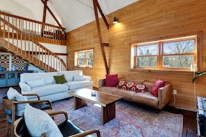 A spacious loft-style living area with a high ceiling, wooden beams, a staircase, and large windows.